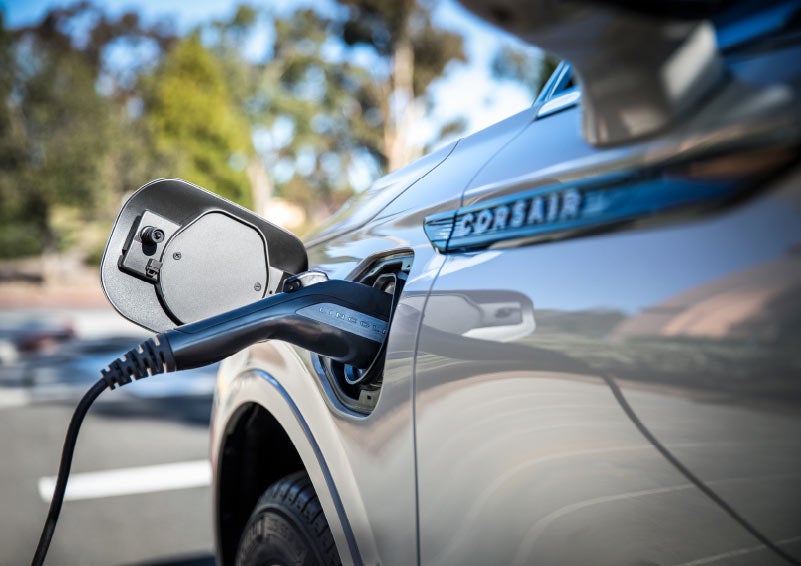 A Lincoln electric charger is plugged into a 2021 Lincoln Corsair Grand Touring port as the body reflects the image of a sunny parking lot