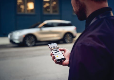 A man is holding his phone looking at the Lincoln Way™ App and we see his 2022 Lincoln Corsair SUV in the background against a blue brick wall | Covert Lincoln Austin in Austin TX